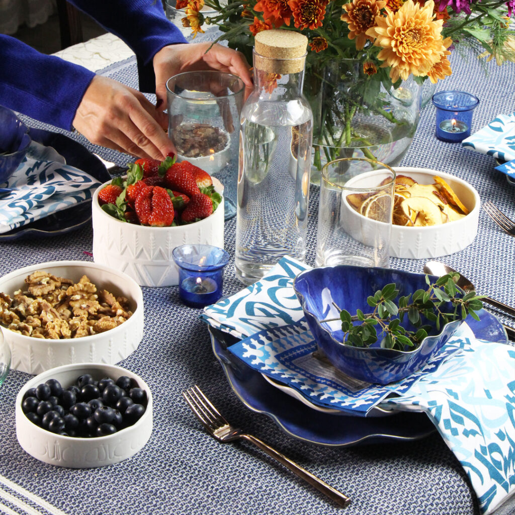 A table topped with plates and bowls of food, including fruit, salad, bread, and meat. The table is set with blue plates and blue napkins.